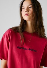 Röd bomullströja med korta ärmar och rundad hals, prydd med "RED BULL RACING" i fet svart text över bröstet.