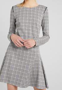 Mujer vestida con un vestido A-line de manga larga y cuadros en blanco y negro, con las manos entrelazadas al frente, sobre un fondo liso gris claro.