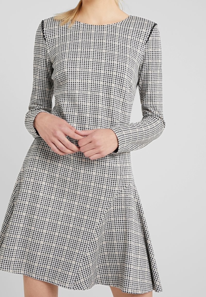 Mujer vestida con un vestido A-line de manga larga y cuadros en blanco y negro, con las manos entrelazadas al frente, sobre un fondo liso gris claro.