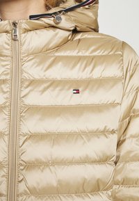 Beige zateplená bunda se zipem a kapucí, s prošívanou texturou a malou nášivkou v červené, bílé a modré barvě na hrudi.