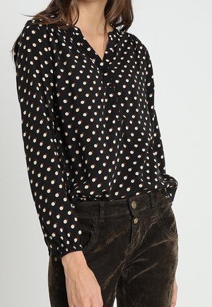 Femme portant un chemisier noir à manches longues à pois blancs et marrons, rentré dans un pantalon en velours côtelé marron foncé.