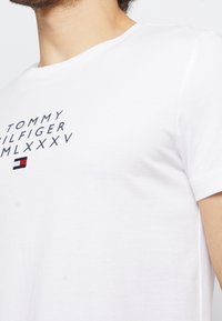Bílý crewneck tričko s textem "Tommy Hilfiger MLXXXV" a malým logem Tommy Hilfiger v červené, bílé a černé barvě na hrudi.