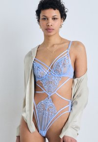 Lingerie in pizzo e rete di un delicato blu presenta motivi floreali, accenti con cinturini e sezioni con ritagli, abbinata a una camicia di colore crema.