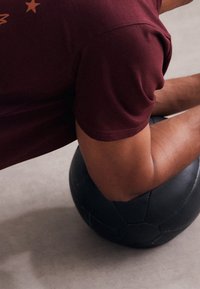 Una pelota de medicina negra sobre un suelo de concreto claro, con una camiseta burdeos con gráficos de estrellas parcialmente visible, mostrando un brazo musculoso en enfoque.