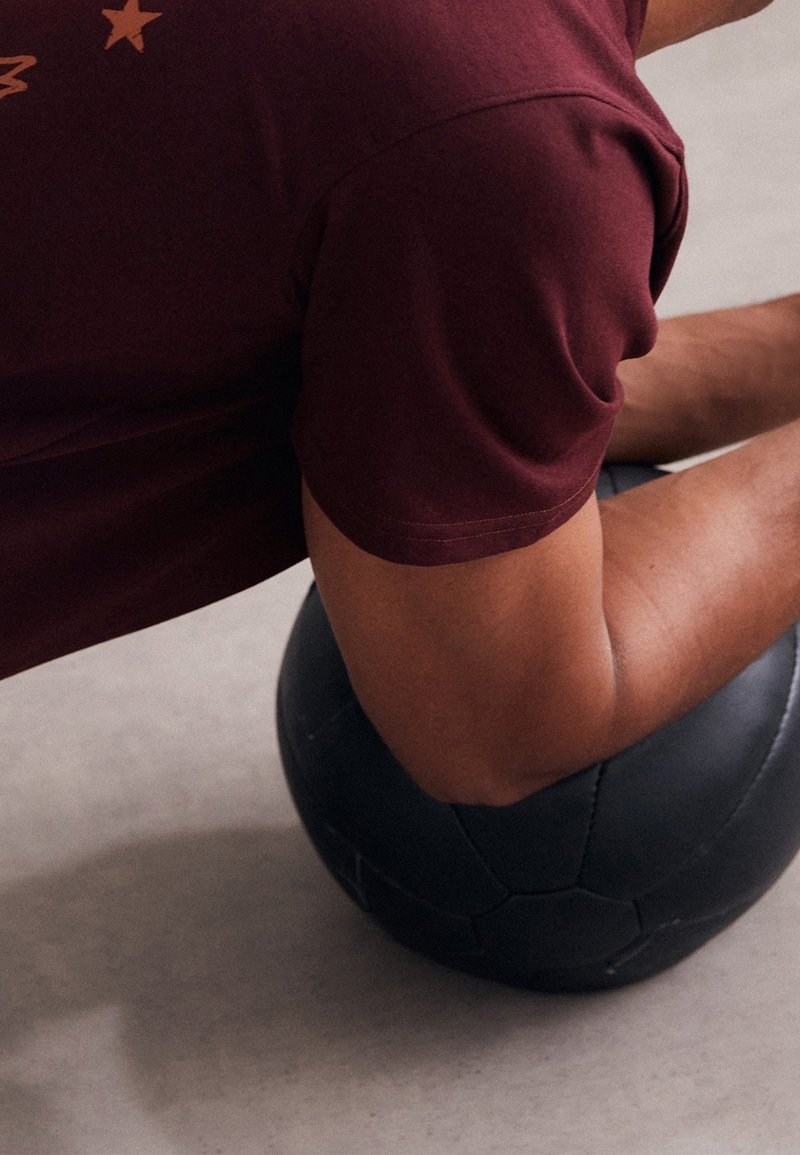 Sort medicinbold på et lyst beton gulv, med en maroon T-shirt med stjerne grafik delvist synlig, der viser en muskuløs arm i fokus.
