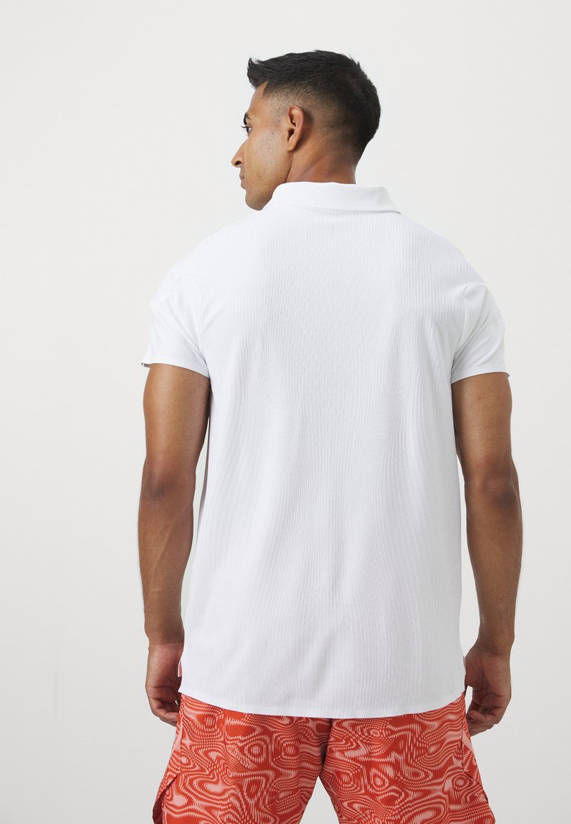 Biała koszulka polo z teksturowanej powierzchni, z krótkimi rękawami i klasycznym kołnierzykiem, zestawiona z jasnopomarańczowymi szortami w wzory.