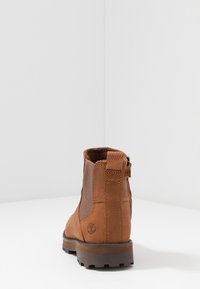 Bottine en cuir marron avec des panneaux latéraux élastiques, une tige texturée et une semelle en caoutchouc. Présente un logo sur le talon et un détail de fermeture éclair.