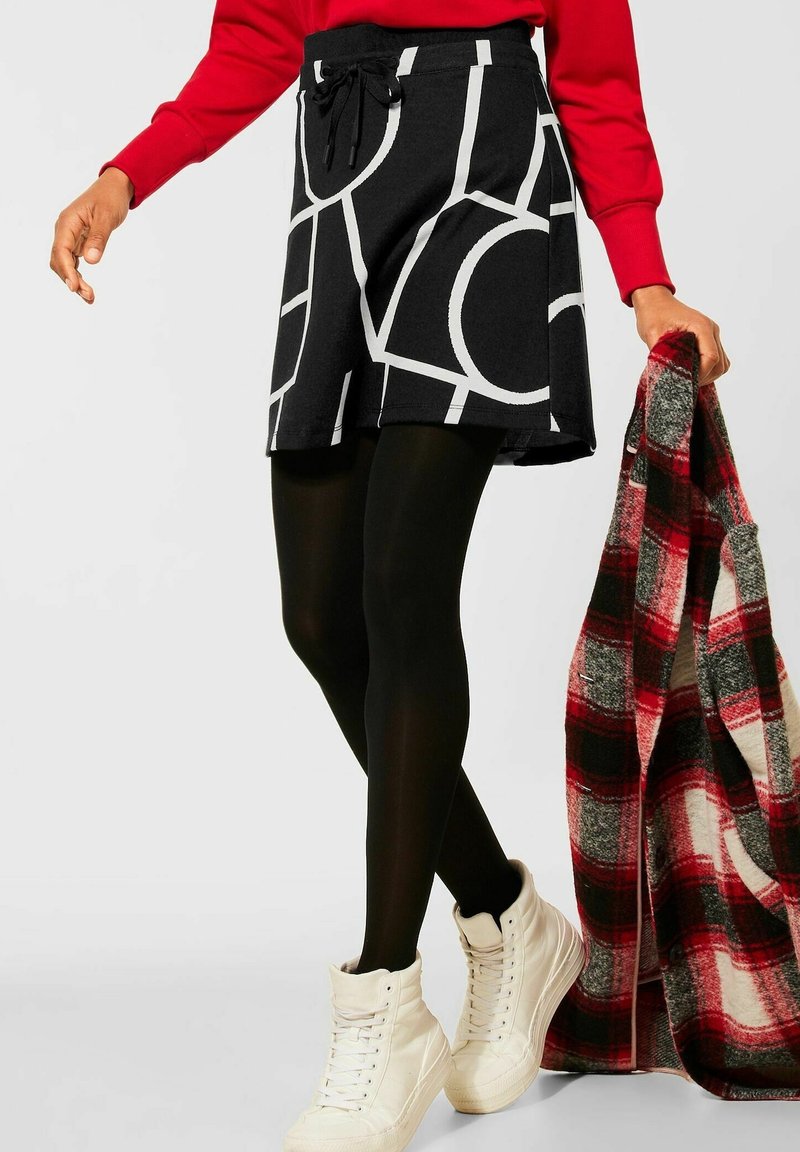 Personne portant une jupe géométrique noire et blanche, des collants noirs, des baskets blanches montantes, un haut à manches longues rouge, tenant un manteau à carreaux rouge et noir.