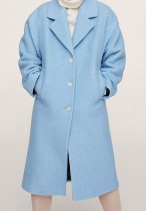 Persona che indossa un cappotto di lana azzurro chiaro con tre bottoni, tasche laterali e un dolcevita color crema sotto, in piedi con le mani nelle tasche.