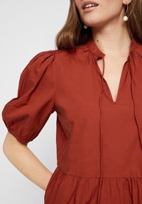 Blusa de color óxido, de manga corta y hecha de tejido ligero. Presenta un escote en V y detalles fruncidos en las mangas.
