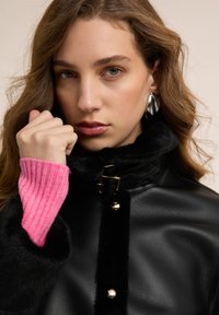 Giovane donna con capelli castani ondulati che indossa orecchini a cerchio d'argento, cappotto in pelle nero con rifinitura in pelliccia e manica di maglione rosa a coste, pugno vicino al viso.