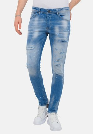 Man draagt slim fit, lichtblauwe distressed jeans met witte sneakers en een wit shirt, staat tegen een effen achtergrond.