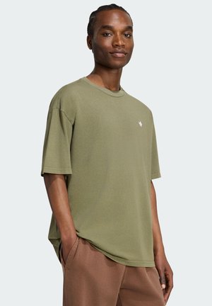 Giovane con capelli intrecciati che indossa una maglietta oversize verde oliva e pantaloni marroni, in piedi con le mani in tasca.