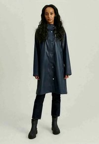 Mujer con cabello largo y negro, vestida con un abrigo de lluvia azul marino oscuro, pantalones negros ajustados y botines negros, de pie contra un fondo simple.