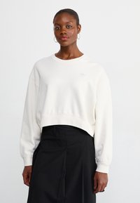 Λευκή κοντή φόρμα με κατεβασμένα μανίκια, ριμπ μανσέτες και ένα μικρό, ανοιχτόχρωμο λογότυπο στο στήθος. Συνδυάζεται με μια σκούρα φούστα.