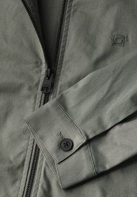 Světlá olivově zelená bunda s černým zipem, detail knoflíku na manžetě a hladkou texturou. Minimalistický design s čistými liniemi.