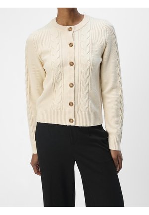 Femme portant un cardigan en maille torsadée crème avec des boutons marron et un pantalon noir, visible du cou aux hanches sur fond blanc.