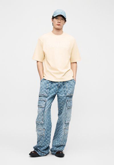 Νεαρός άνδρας που φοράει ανοιχτό μπεζ φαρδιά μπλούζα, μπλε cargo τζιν με σχέδια, τζιν καπέλο και μαύρα παπούτσια, στέκεται με τα χέρια στις τσέπες.