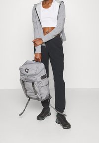 Persona con una sudadera gris, top corto blanco, pantalones negros, zapatillas negras, sosteniendo una mochila gris de Burton por su asa.