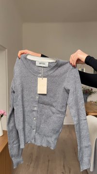 Grauer Cardigan mit rundem Halsausschnitt, langen Ärmeln, fünf Knöpfen vorne und leichter Struktur. Markenzeichen angebracht.