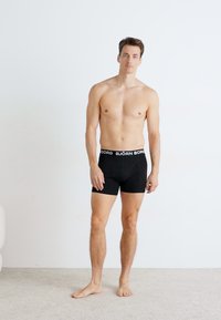 Schwarze Boxershorts mit einem weißen "BJÖRN BORG" Bund, glattes Material, figurbetontes Design, Modell steht auf einem hellen Teppich vor einer schlichten Wand.