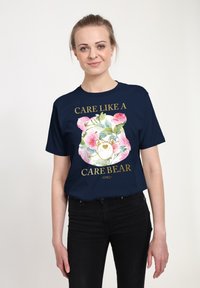 Marinblå t-shirt med en blommig Care Bear-grafik, som har rosa rosor och guldtexten "CARE LIKE A CARE BEAR" med året 1982.