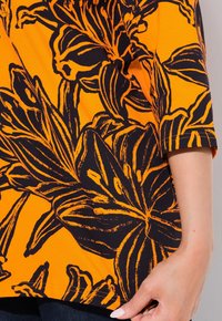 Orange T-Shirt mit einem schwarzen Blumenmuster. Kurze Ärmel, weicher Stoff und eine lockere Passform betonen die auffälligen Blumenmuster und Linien.