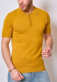 Polo à manches courtes en jaune moutarde, fabriqué en tissu lisse. Il présente un col et une patte de boutonnage à trois boutons, avec des poignets côtelés.