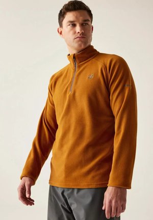 Hombre con un pulóver de forro polar color óxido con cremallera hasta un cuarto, de pie y mirando de lado contra un fondo liso.