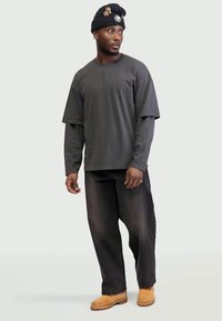 Man wearing layered dark gray long-sleeve and short-sleeve shirts, black pants, tan boots, and a black beanie with pins, looking to the side.