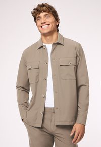 Beige Hemd mit Knöpfen, zwei Brusttaschen, langen Ärmeln und einem spitzen Kragen, kombiniert mit passenden beigen Hosen. Glatter Stoff.