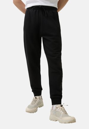Zwarte sweatpants met een trekkoord in de taille, taps toelopende benen en ribbels aan de boorden. Draagt het samen met lichtgekleurde sneakers met een dikke zool.
