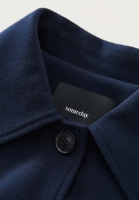 Navy blue jacket with a collared neckline, featuring a black button, textured fabric, and a label displaying "someday."