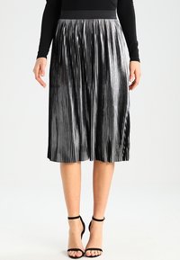 Falda negra plisada con un acabado metálico brillante. Diseño de longitud media con rayas verticales. Combinada con zapatos de tacón negros con tiras.