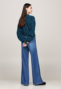 Suéter texturizado en color azul verdoso con mangas abullonadas, combinado con pantalones de mezclilla de pierna ancha y cintura alta en azul. Zapatos negros completan el conjunto. Vista trasera.