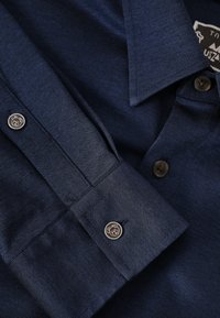 Tumši zila pogājamā krekla ar teksturētu audumu, ar norādītu apkakli un dekoratīvām metāla pogām uz piedurknēm.