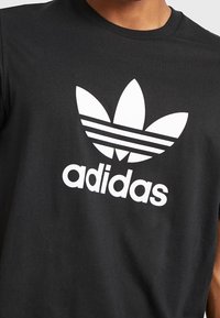 Černé bavlněné tričko se velkým bílým logem Adidas a třemi pruhy nad ním. Tkanina vypadá hladce a má design s kulatým výstřihem.