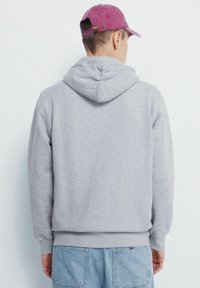 Szara bluza z kapturem, wykonana z miękkiego materiału, z kieszenią kangurką, ściągaczami na rękawach i luźnym krojem. Widać jasnoniebieskie dżinsy.