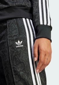 Čierny športový tréningový oblek s textúrovaným vzorom, biele logo adidas a tri biele pruhy pozdĺž rukávov a strán nohavíc.