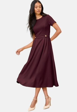 Ein burgunderrotes Kleid mit tailliertem Oberteil, kurzen Ärmeln und plissiertem Rock. Akzentknöpfe an der Taille. Getragen mit beigen High-Heels.