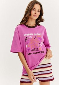 Růžové oversized tričko s grafickým potiskem a hnědým výstřihem, zkombinované s pletenými šortkami s vícero barevnými pruhy. Měkká tkanina, volný střih.