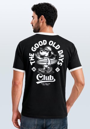 Mann trägt schwarzes T-Shirt mit weißem Rand, zeigt Cartoon-Hund mit Surfbrett und Text "The Good Old Days Club" auf dem Rücken.