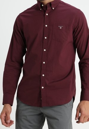 Mężczyzna w ciemnoczerwonej, zapinanej na guziki koszuli z długim rękawem i kieszenią na piersi oraz szarych spodniach na jednolitym tle.
