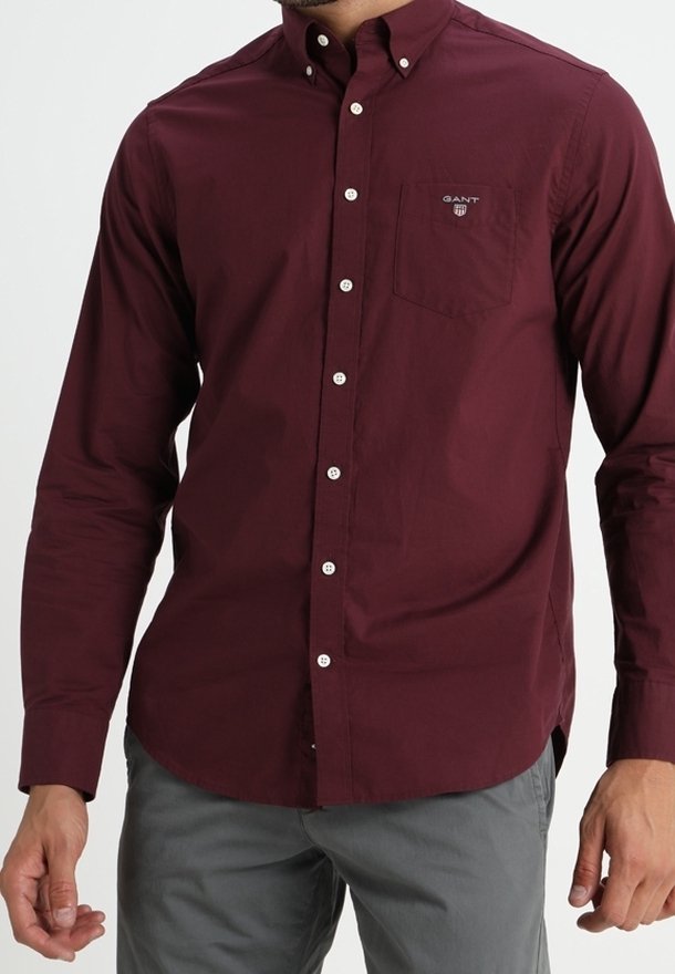 Homme portant une chemise rouge foncé à manches longues boutonnée avec une poche poitrine et un pantalon gris sur un fond uni.