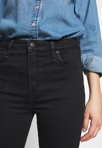 Jeans de mezclilla negra de cintura alta, con diseño de cinco bolsillos y botón de metal plateado. Llevados con una camisa de mezclilla azul.