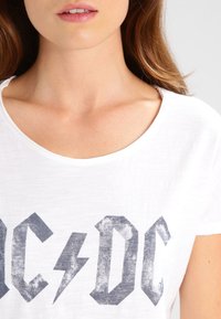 Camiseta de algodón blanca con cuello redondo, que presenta un gráfico en negro desgastado de AC/DC con letras en negrita en el pecho.