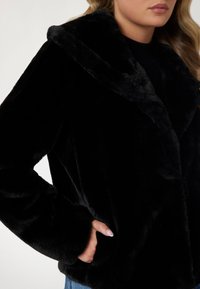 Veste en fausse fourrure noire avec col châle, manches longues et poches latérales. Texture douce et aspect moelleux pour plus de chaleur et de confort.