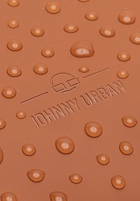 Saltea de cauciuc maro cu picături de apă, logo-ul „JOHNNY URBAN” embosat, prezintă o suprafață texturată și un model circular creat de picături.