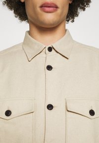 Beżowa koszula z zapięciem na guziki, z kołnierzykiem, posiadająca dwie kieszenie na piersi i czarne guziki. Faktura wydaje się lekko chropowata i trwała.