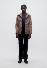 Ενήλικας που φοράει καφετί μπουφάν με κουκούλα, μαύρο παντελόνι και μαύρα παπούτσια, στέκεται με το σώμα προς τα εμπρός μπροστά σε ένα απλό φωτεινό φόντο.